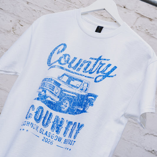 The C2C Country To Country Pick Up White T-Shirt, a unisex tailored fit, hangs on a white brick wall. It features a blue vintage pickup truck graphic and "Country, Country, London, Glasgow, Belfast 2026" text.