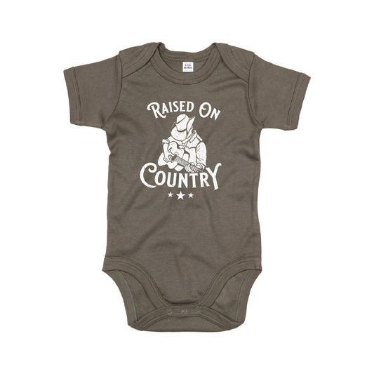 The C2C Country To Country Store Baby Grow is an olive green onesie with a white graphic of a cowboy playing guitar, the text Raised On Country, and three stars beneath the image.