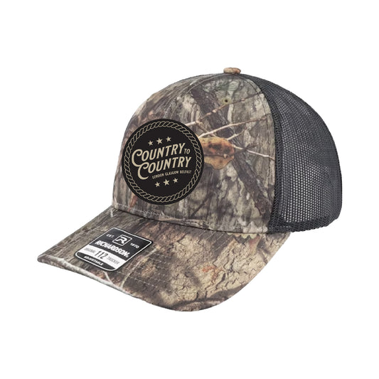 The Camo Cap from C2C Country To Country Store features a camouflage design, black mesh back, and a circular patch with yellow Country to Country text—tags attached to the brim complete this rugged, stylish accessory.
