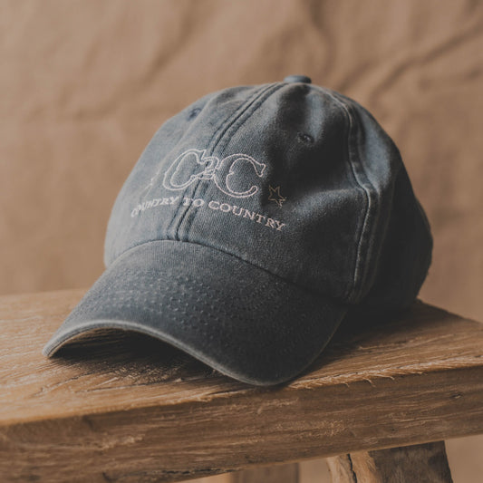 The C2C Country To Country Store Dad Cap, a faded black baseball cap with C2C and Country to Country embroidered on the front, is perfect for fans of the country music festival. Displayed on rustic wood against a brown background.
