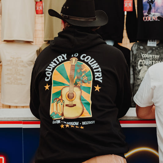 A person in a black cowboy hat and a C2C Country To Country Store PRE-ORDER: 2026 Online Exclusive Hoodie with guitar and flower designs stands at a counter beside someone in a white shirt.