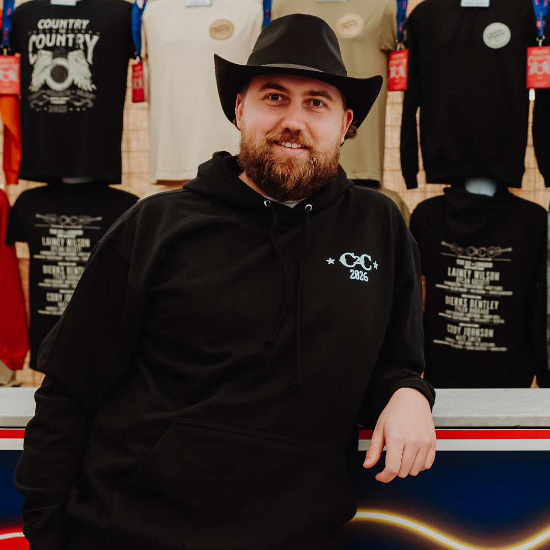 A bearded man in a black cowboy hat and hoodie smiles by country-themed t-shirts, including the C2C Country To Country Store's PRE-ORDER: 2026 Online Exclusive Hoodie with front and back print.