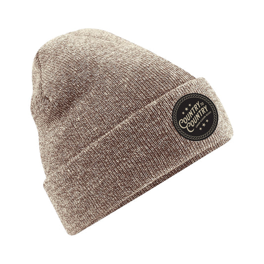The Heather Oatmeal Beanie Hat from C2C Country To Country Store features a folded brim and a black circular patch reading “Country by Country.” Displayed on white, this warm knit accessory is perfect for winter outfits.