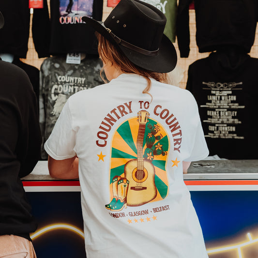 A person in a black cowboy hat and a white PRE-ORDER: 2026 Online Exclusive T-Shirt from the C2C Country To Country Store stands at a merchandise counter, with more exclusive event t-shirts displayed in the background.