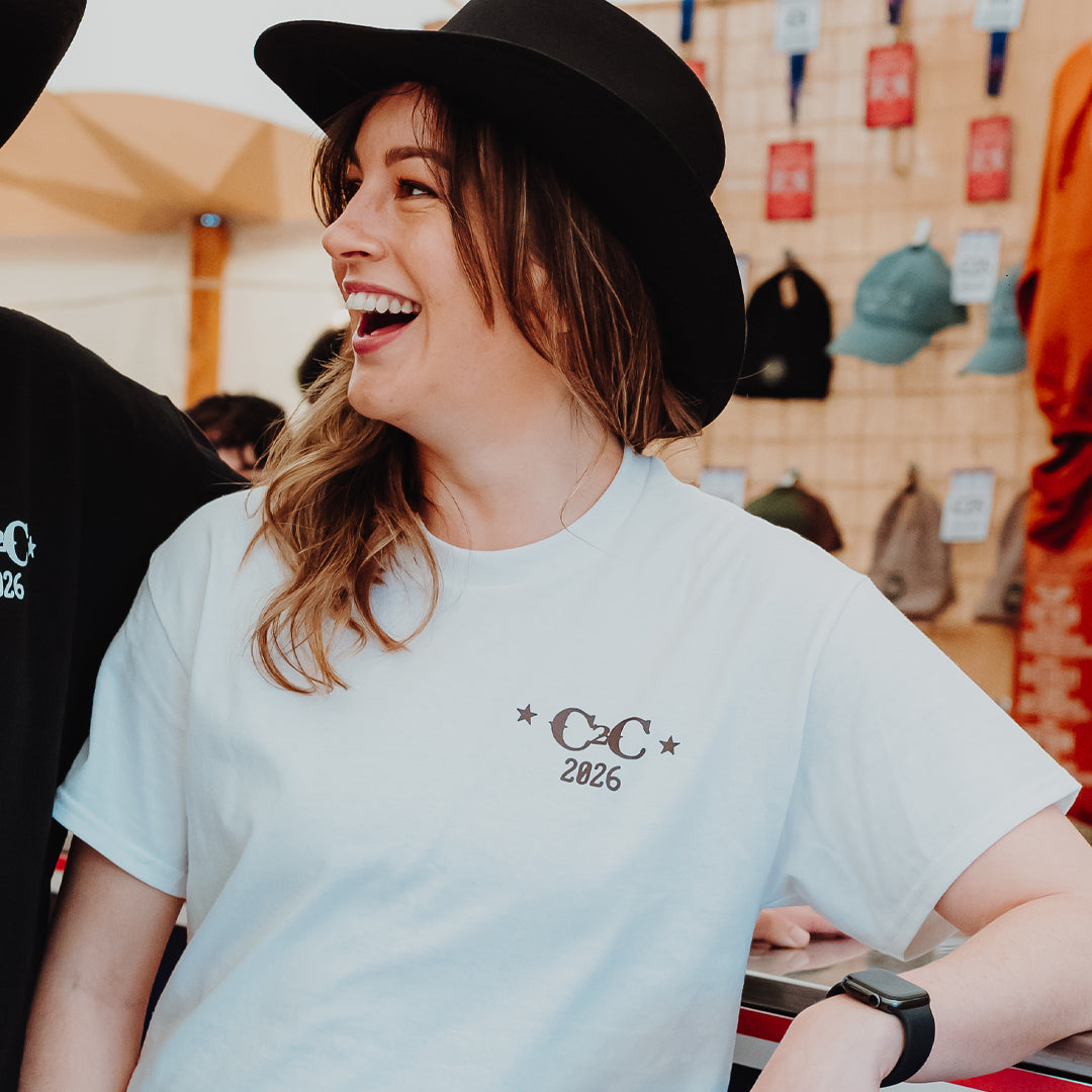 A person with long hair in a black wide-brim hat smiles while wearing the C2C Country To Country Store PRE-ORDER: 2026 Online Exclusive T-Shirt. Hats and other items appear on the wall in the background.