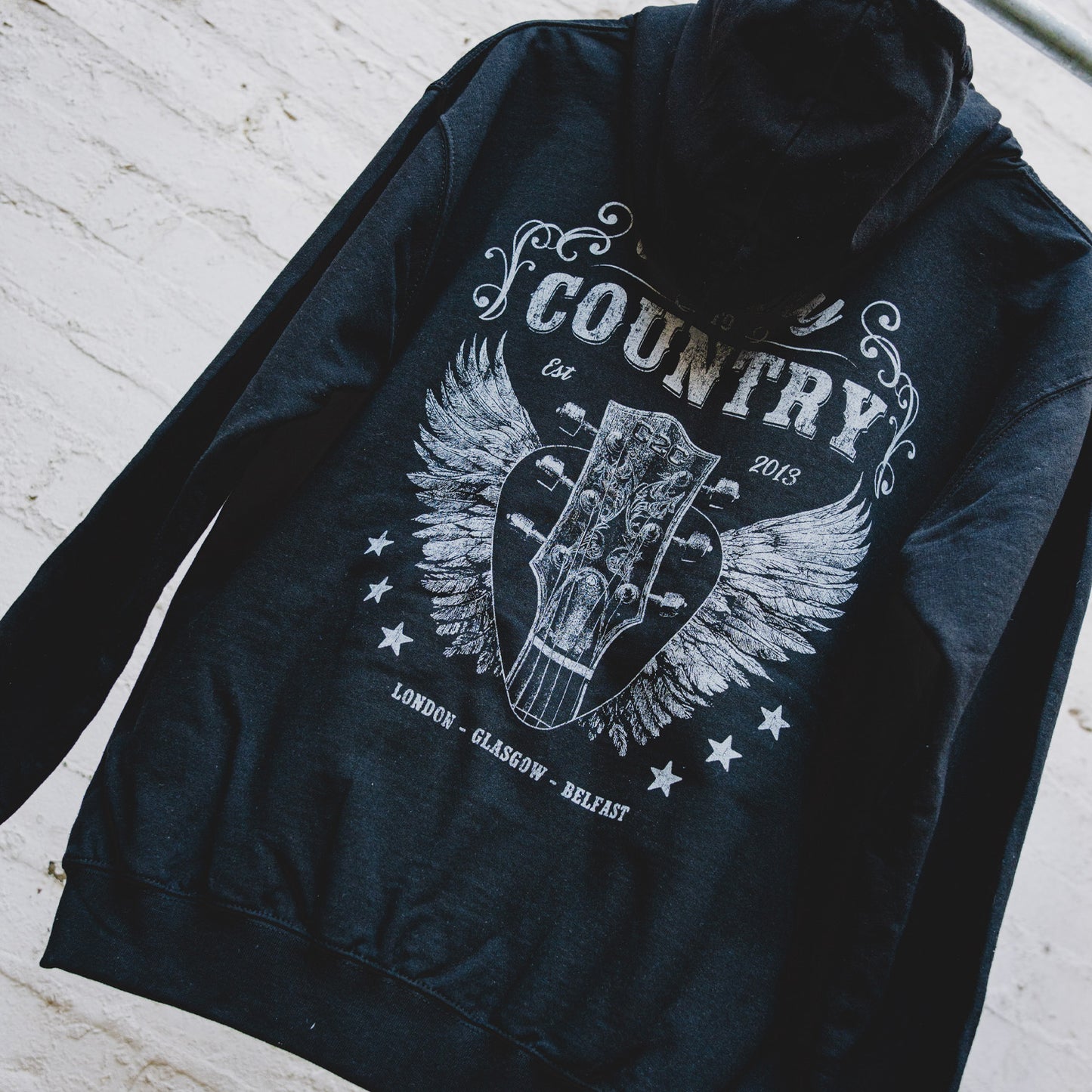 The C2C Country To Country Angel Guitar Black Zip Hoodie, made from soft cotton with a relaxed fit, features a bold guitar headstock graphic with wings, stars, and "Country, Est 2013, London - Glasgow - Belfast" text.