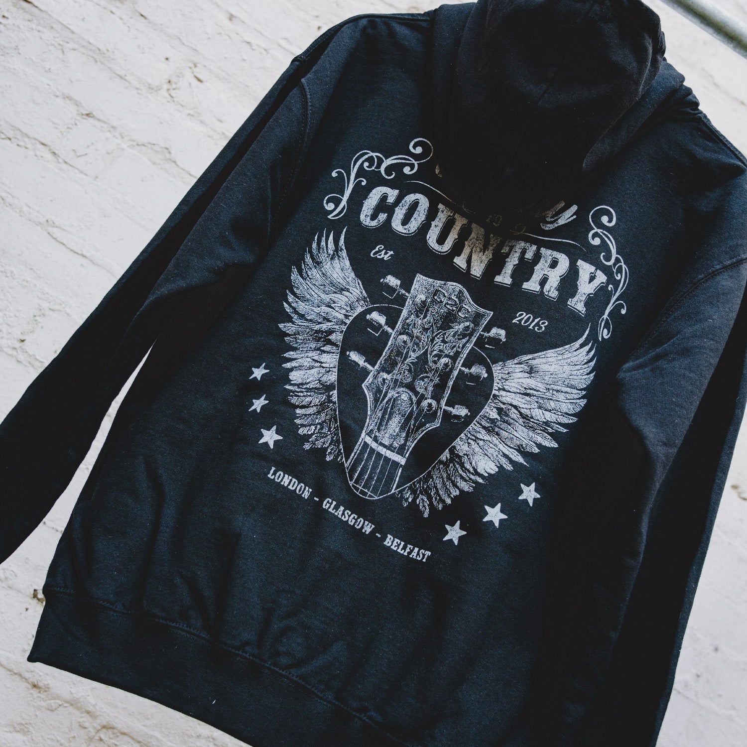 The C2C Country To Country Angel Guitar Black Zip Hoodie, made from soft cotton with a relaxed fit, features a bold guitar headstock graphic with wings, stars, and "Country, Est 2013, London - Glasgow - Belfast" text.
