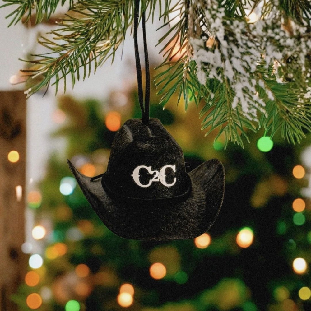 The C2C Black Cowboy Hat Bauble by C2C Country To Country, featuring a silver buckle, hangs from a snow-dusted pine branch with blurred colorful holiday lights in the background.