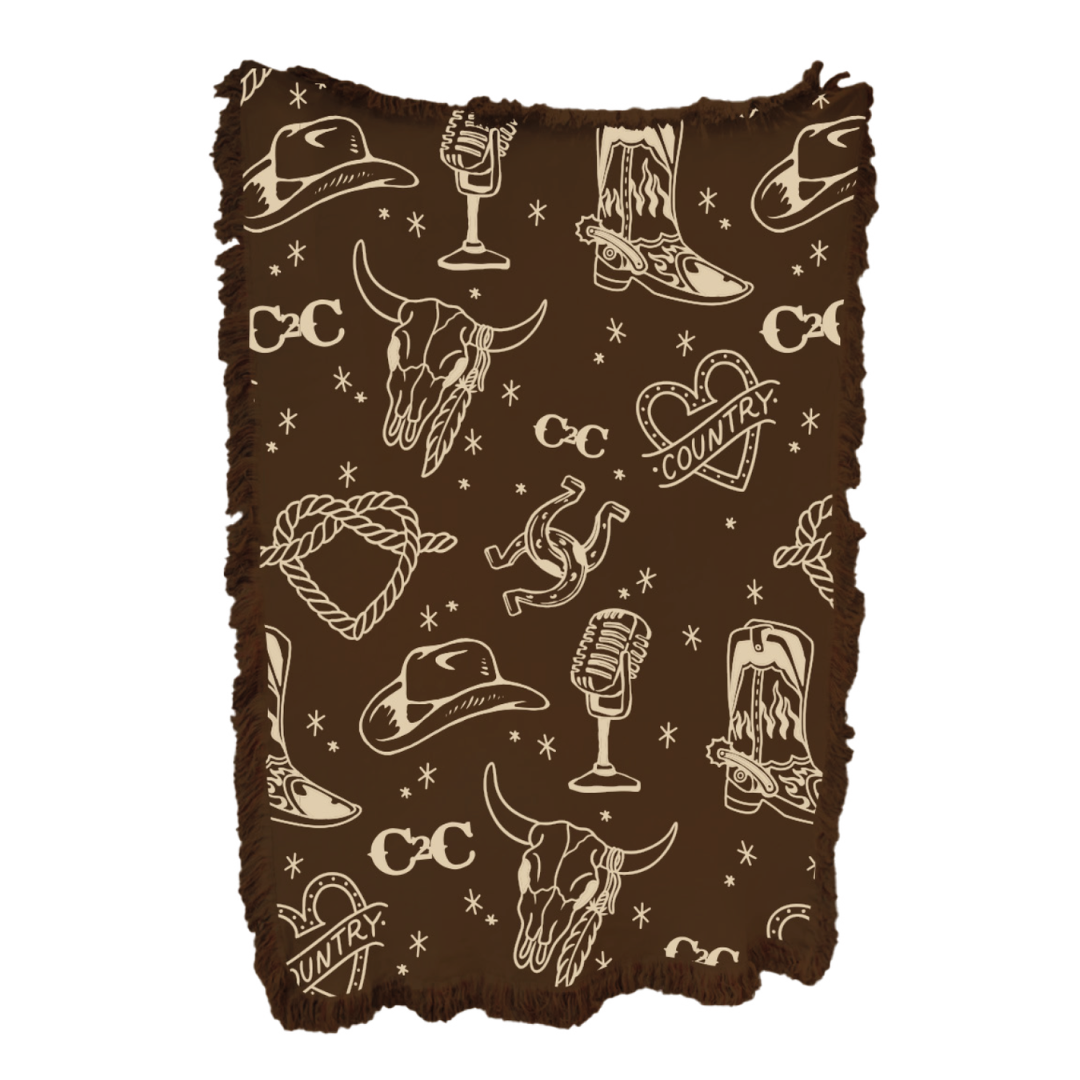 The 2026 Blanket features brown fringe and a cream pattern with cowboy hats, boots, bull skulls, lassos, microphones, horseshoes, snakes, hearts labeled "Country," and stars—a cozy piece for country music-inspired decor.