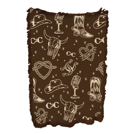 The 2026 Blanket features brown fringe and a cream pattern with cowboy hats, boots, bull skulls, lassos, microphones, horseshoes, snakes, hearts labeled "Country," and stars—a cozy piece for country music-inspired decor.